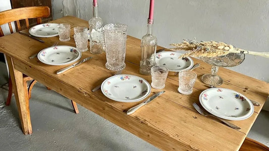 Table de ferme en bois clair dressée avec de la vaisselle ancienne : assiettes blanches à motifs floraux, verres et carafes en verre ciselé, et couverts vintage. Un bouquet séché sur une coupe en verre décore le centre devant un mur épuré.