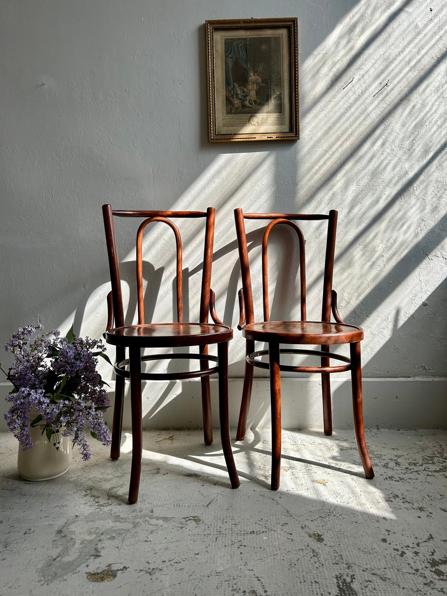 Paire de chaises bistrot anciennes en bois courbé de style Thonet, placées côte à côte contre un mur blanc baigné de lumière. À gauche, un pot en grès garni de branches de lilas violet. Un cadre ancien doré est suspendu au-dessus des chaises.