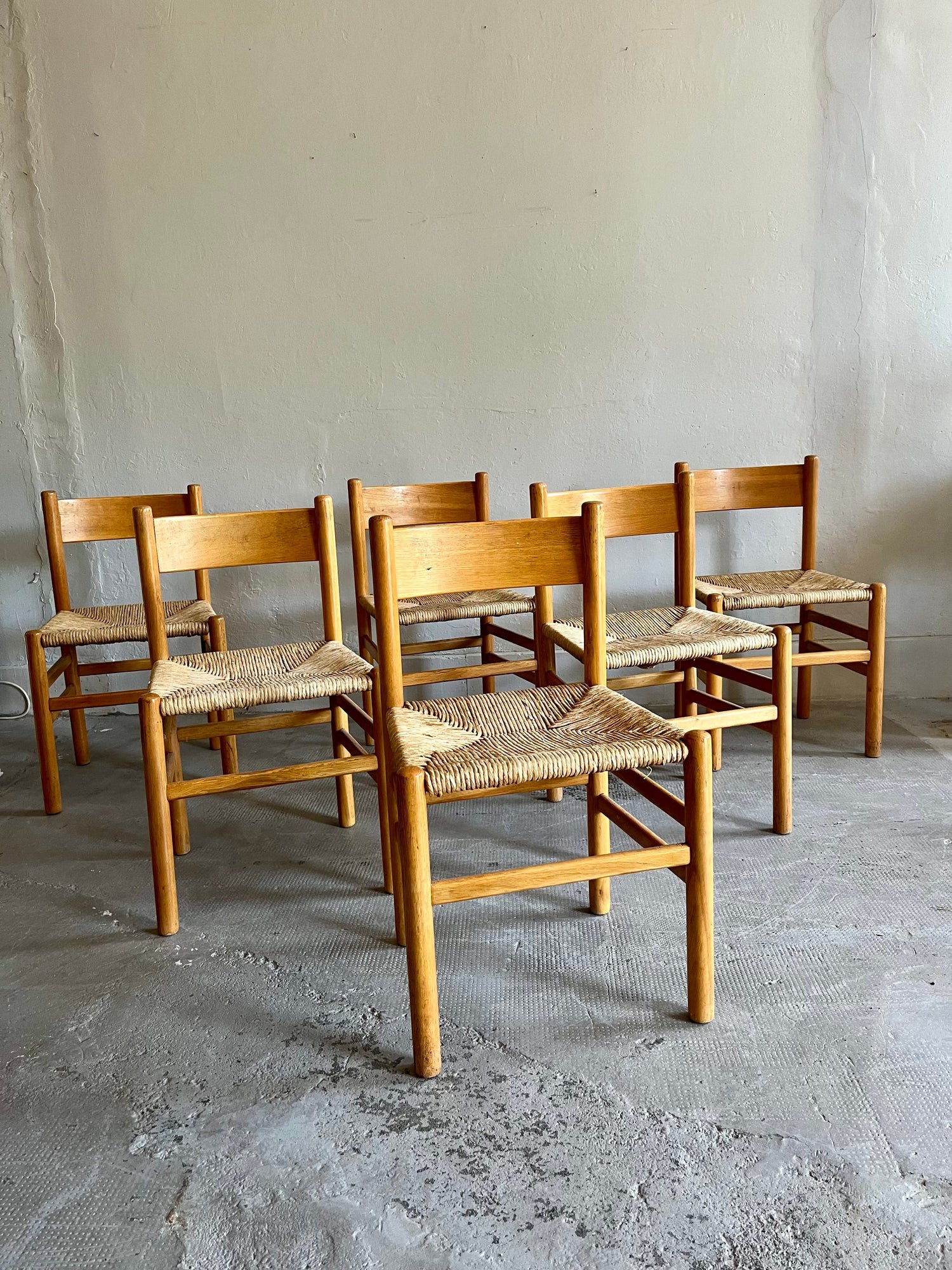 Série de six chaises vintage de style brutaliste en bois clair (chêne ou hêtre) avec assise paillée en corde tressée. Disposées en groupe dans une pièce au sol en béton brut et mur gris clair.