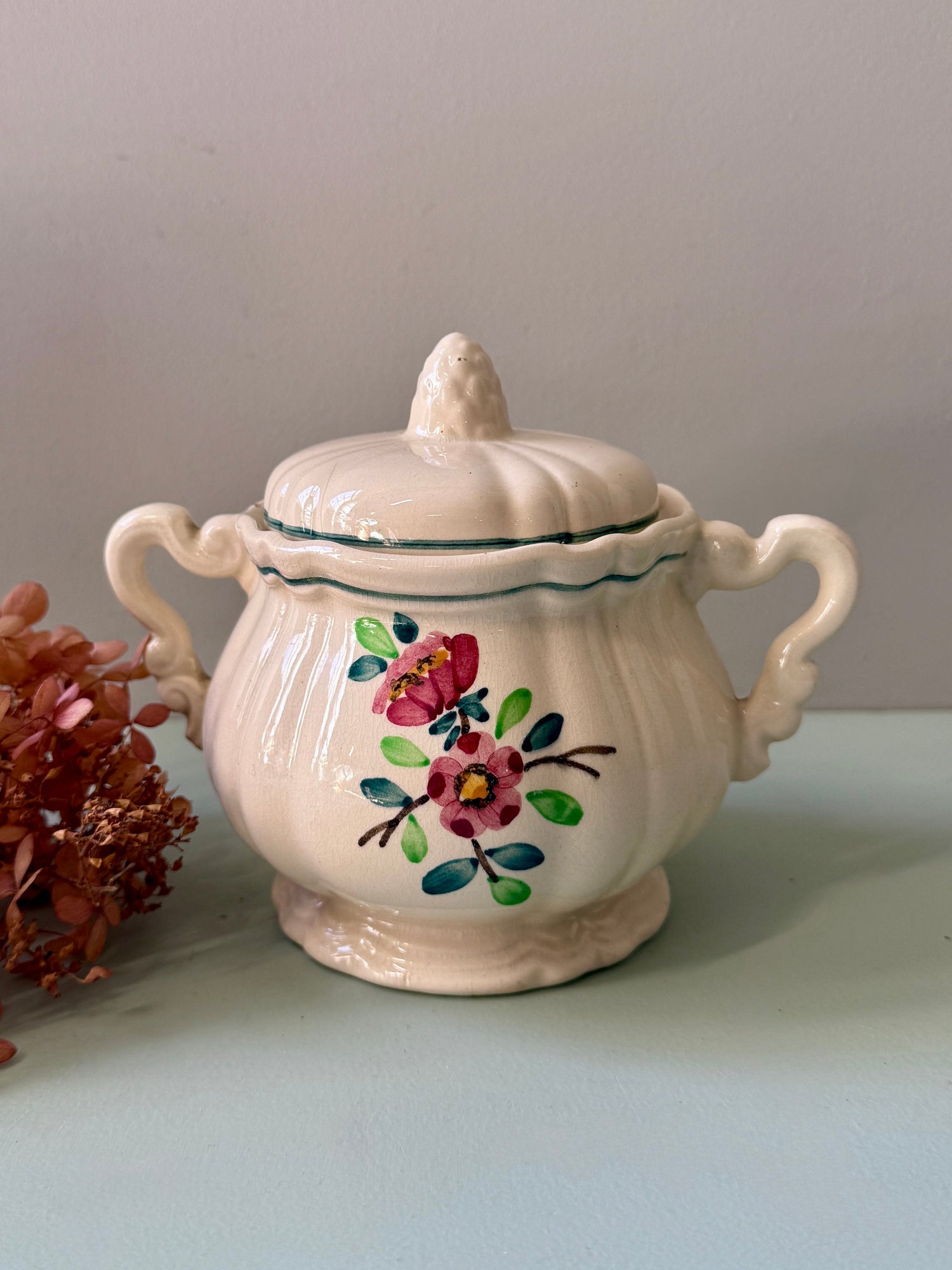Sucrier blanc avec deux anses sur un fond vert clair. Décor floral teintes rose et vert. Une fleur posée à côté