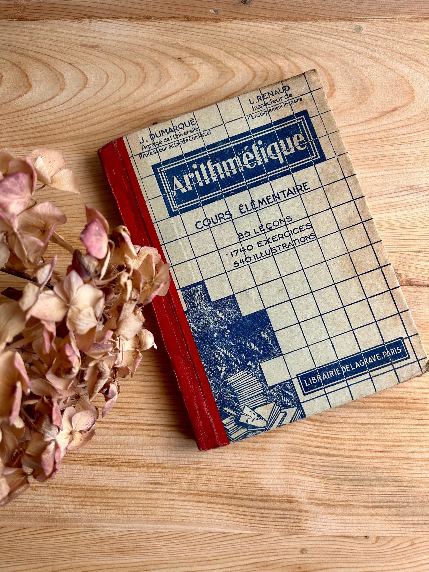 Livre scolaire ancien d'arithmétique pour cours élémentaire par J. Dumarqué et L. Renaud, Éditions Librairie Delagrave. Couverture rétro quadrillée avec tranche rouge, posé à plat sur une table en bois clair à côté d'hortensias séchés.