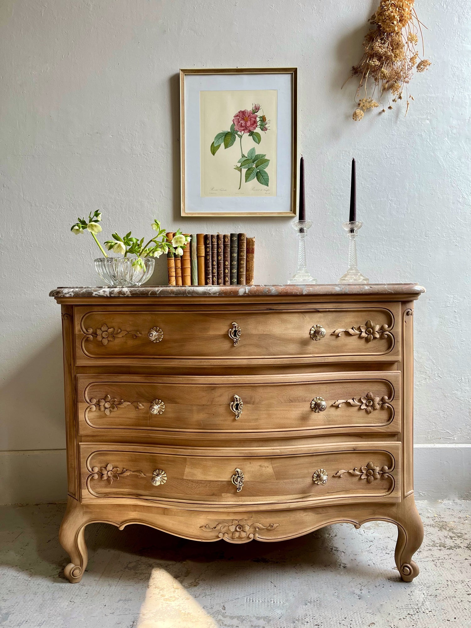 Commode ancienne en bois brut de style Louis XV avec trois tiroirs sculptés et plateau en marbre, décorée de livres anciens et d'une gravure botanique.