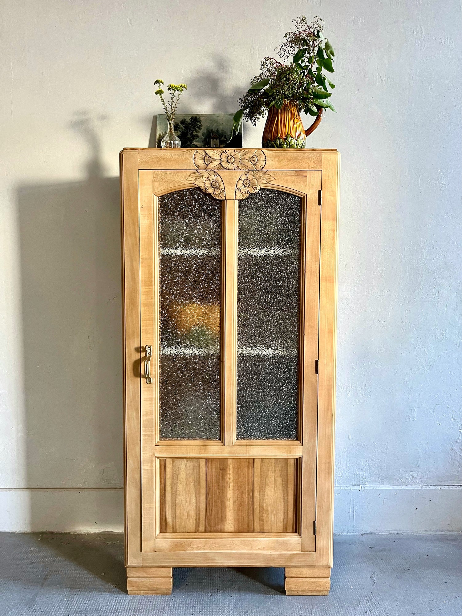Armoire vitrée vintage en bois clair sculpté de motifs floraux en haut de porte, avec vitres en verre texturé flou. Le meuble est surmonté de fleurs dans de petits vases et posé contre un mur clair.