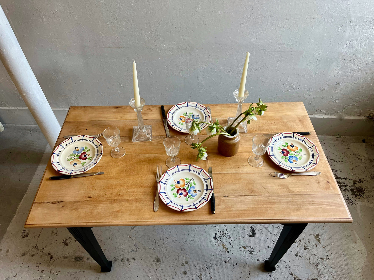 Vue du dessus table bistrot en bois dressee avec assiette ancienne, couverts en argent, verre en cristal. 2 bougeoirs en verres ciselé, un pot à moutarde avec des fleurs.