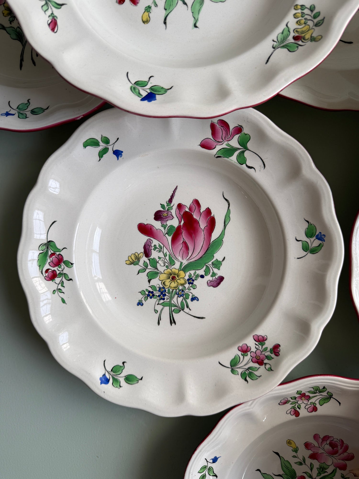 Assiette creuse ancienne en faïence blanche à bord festonné et liseré bordeaux, décorée au centre d'un bouquet peint de tulipe et fleurs multicolores (rose, jaune, bleu). Présentée à plat parmi d'autres assiettes assorties.