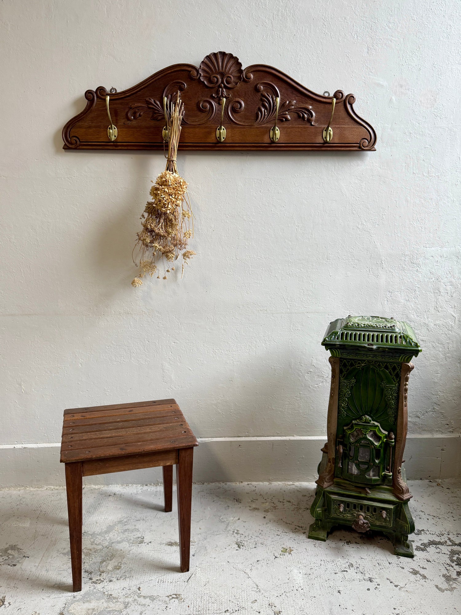 Grand porte-manteau mural sculpté en bois foncé avec crochets dorés, d'où pend un bouquet séché. Un tabouret en bois lattes à gauche et un petit poêle en fonte émaillée verte complètent ce décor rétro sur mur blanc.