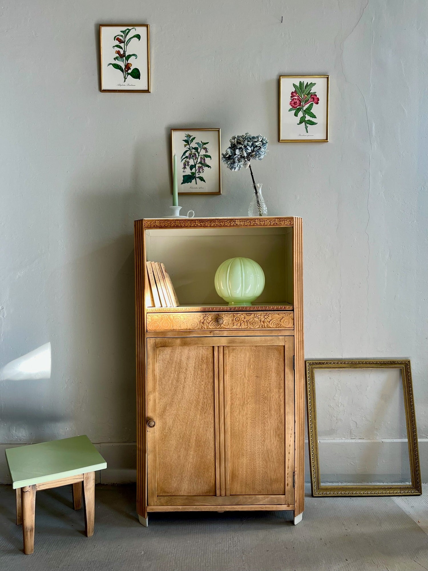 Meuble de rangement vintage en bois clair avec une niche ouverte et une porte basse, mis en scène dans un décor avec une lampe globe vert pastel, un petit tabouret assorti et des cadres botaniques anciens sur un mur gris.