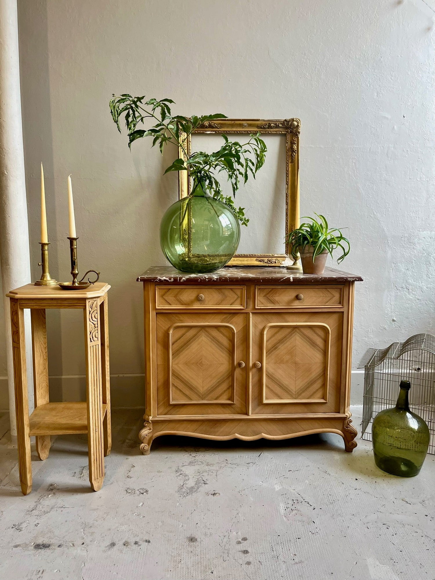 Mise en scène d'un buffet ancien en bois clair avec plateau en marbre, accompagné d'une sellette assortie, d'une grande dame-jeanne en verre vert et d'un cadre doré vide sur un mur gris épuré.