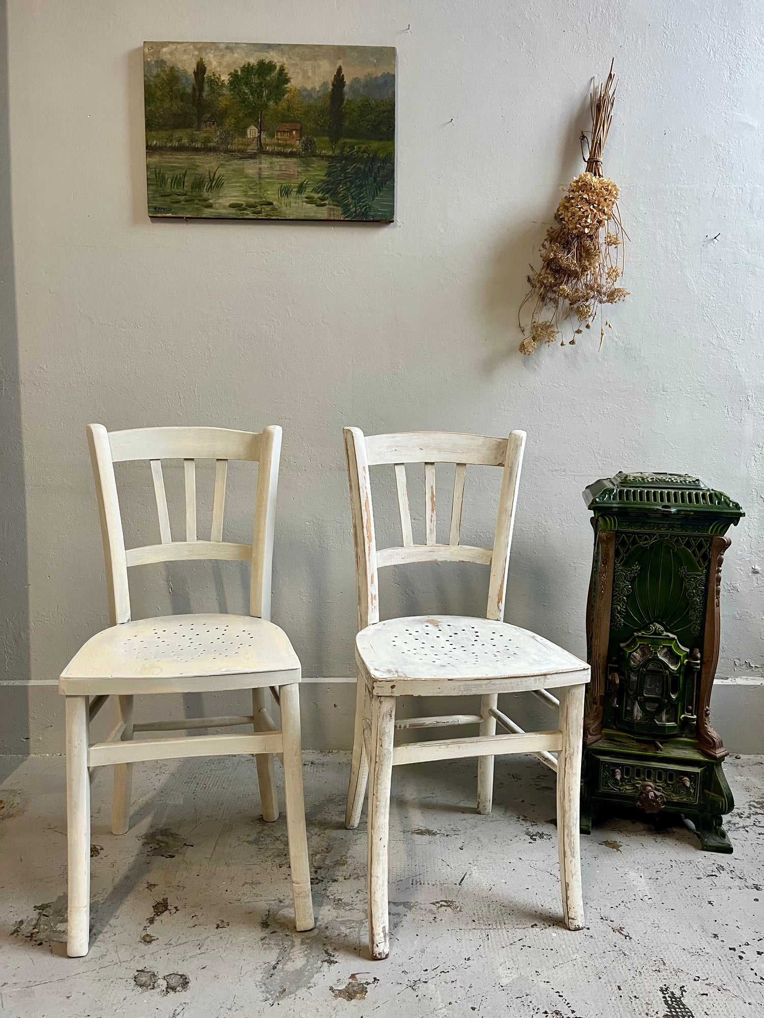 Paire de chaises bistrot en bois patiné blanc, assises perforées. À droite, petit poêle ancien en fonte émaillée verte. Au mur gris clair, un tableau de paysage bucolique et un bouquet de fleurs séchées suspendu complètent cette scène rétro.