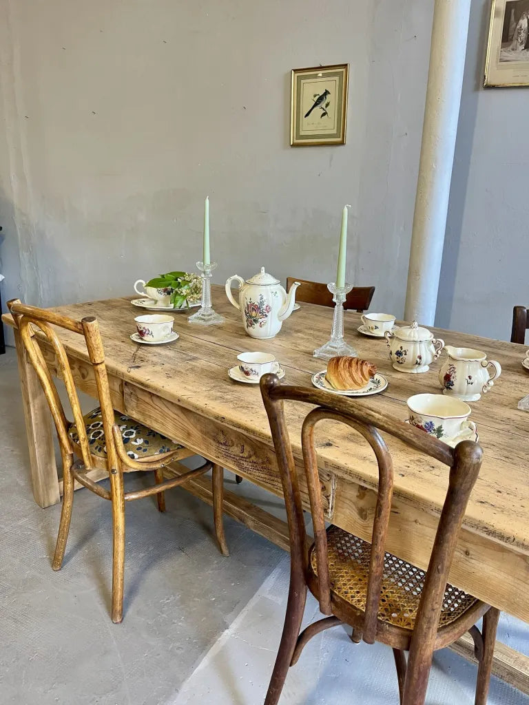 Grande table de ferme ancienne en bois brut entourée de chaises dépareillées rétro. Dessus dressé : vaisselle fleurie en faïence avec théière, tasses, pot à lait, sucrier et un croissant, deux bougeoirs en verre avec bougies vertes. Cadres vintage au mur.