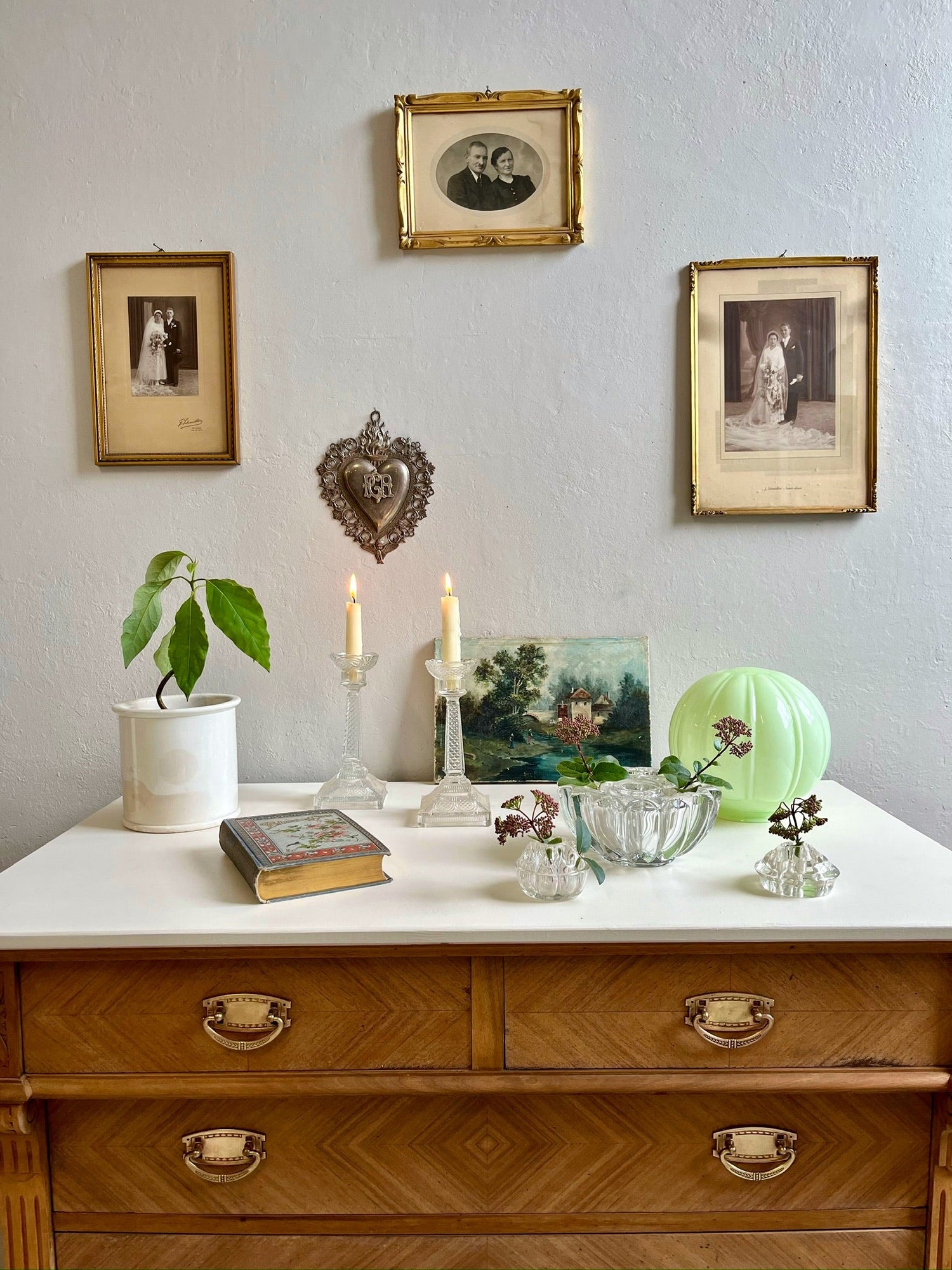 Mise en scène d'objets de décoration vintage sur une commode en bois clair, incluant des bougeoirs en verre, un ex-voto en métal, une lampe globe vert pastel et des cadres anciens avec photographies de mariage en noir et blanc sur un mur gris.