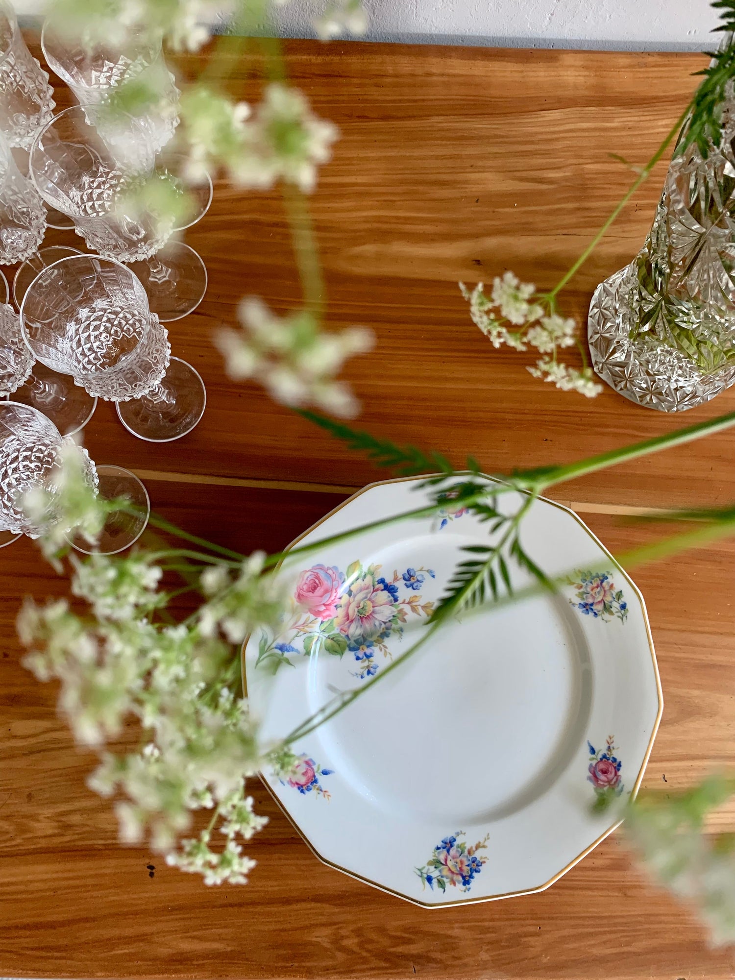 Assiette vintage en porcelaine blanche aux motifs floraux et bordure dorée, posée sur une table en bois clair à côté de verres en cristal gravé et de branches de fleurs blanches.