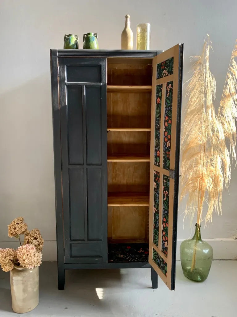 Armoire parisienne ancienne restaurée et peinte en noir ou bleu foncé, présentée avec une porte ouverte révélant des étagères en bois brut et un intérieur de porte tapissé de tissu fleuri, accompagnée d'une grande dame-jeanne avec des plumes de pampa.