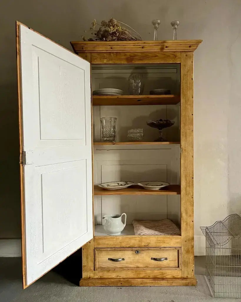 Armoire bonnetière ancienne en bois clair avec porte ouverte peinte en blanc à l'intérieur, dévoilant des étagères garnies de vaisselle (assiettes, verrerie, broc blanc). Tiroir en bas, fleurs séchées au sommet, posée contre un mur clair.