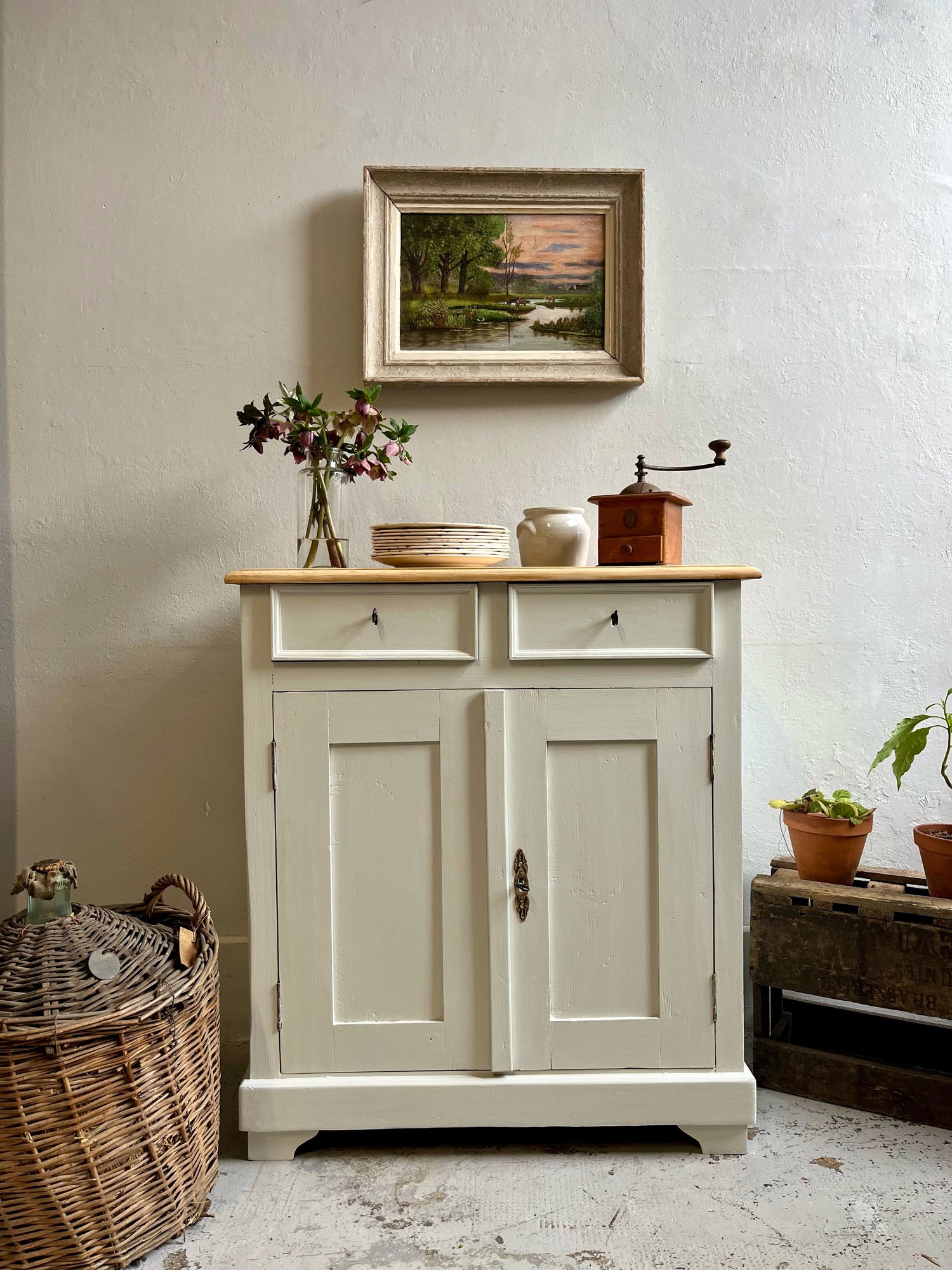 Buffet parisien ancien peint en beige clair avec plateau en bois brut. Dessus posés : vase de fleurs, pile d'assiettes, pot en grès et moulin à café en bois. Au mur, un tableau paysage encadré. À côté, un grand panier en osier et une caisse.