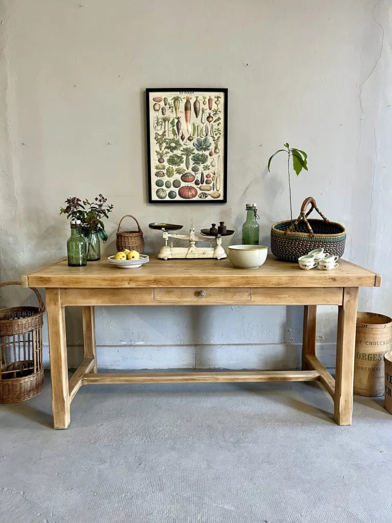 etabli ancien avec tiroir Grande table de ferme ancienne en bois clair avec tiroir central, mise en scène avec une balance de cuisine vintage, des paniers en osier et une affiche botanique sur un mur gris dans un style brocante chic.