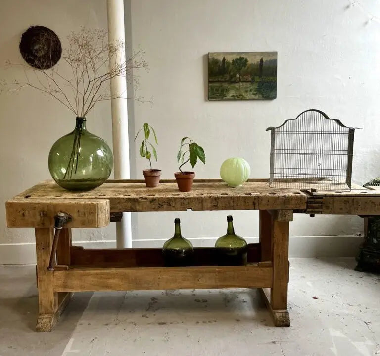 Ancien établi de menuisier en bois brut avec sa presse. Dessus : grande dame-jeanne verte avec branchages, deux petits pots en terre cuite, un globe en opaline verte et une cage à oiseaux en métal. Deux dames-jeannes sont posées en bas.