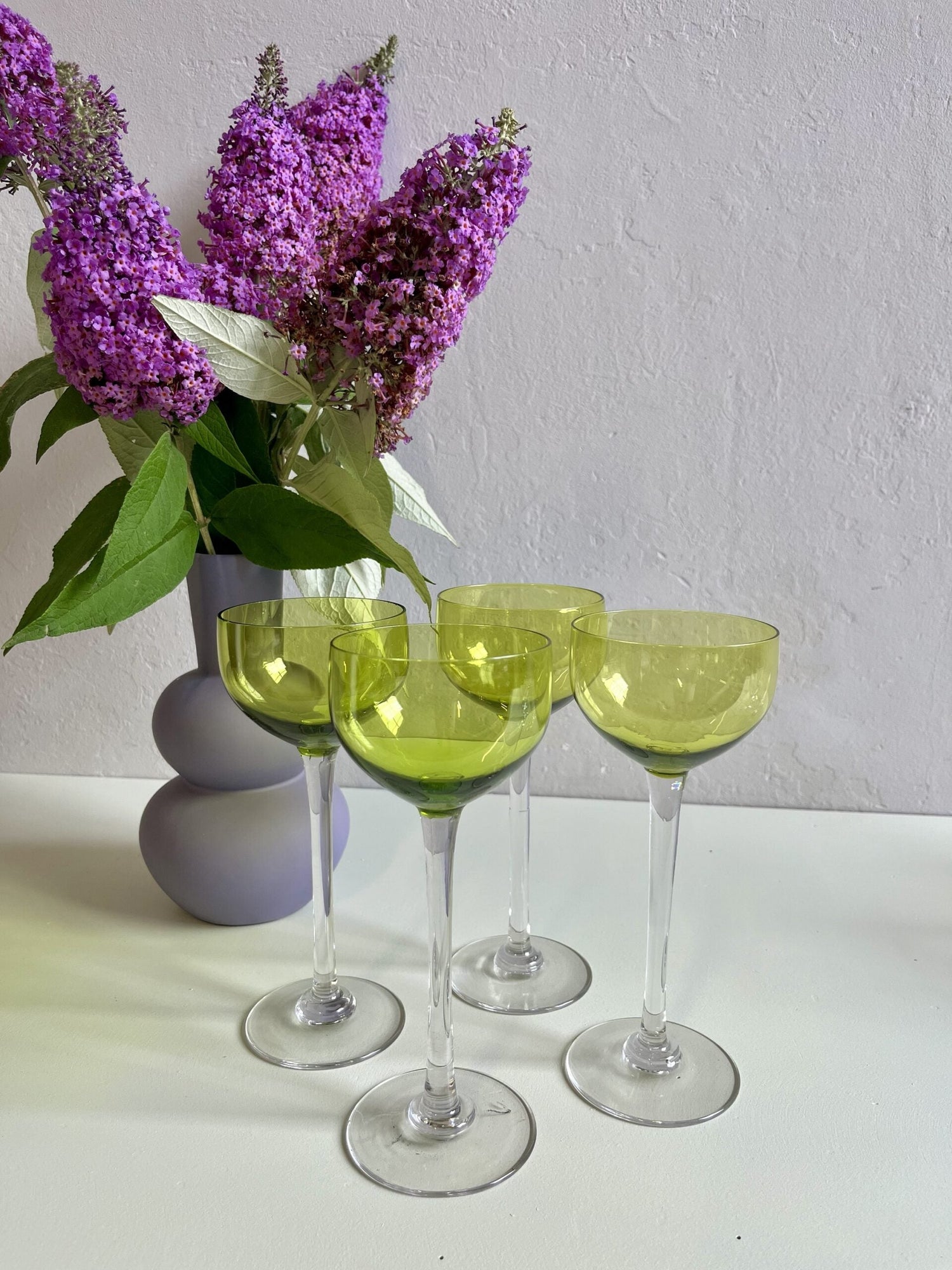 Ensemble de quatre verres à pied vintage au calice teinté d'un vert olive translucide et à la jambe transparente. À l'arrière-plan, un vase lilas de forme organique accueille un bouquet de buddleia (arbre aux papillons) violet vif contre un mur blanc.
