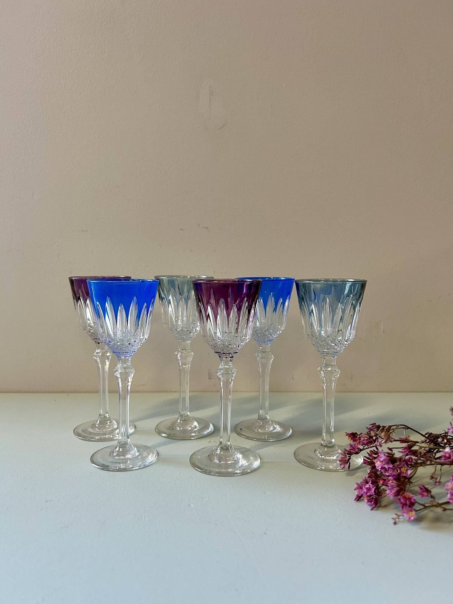 Série de six verres en cristal taillé doublé de couleur (bleu, violet, turquoise) sur fond clair. À droite, de petites fleurs séchées violettes complètent cette élégante composition vintage.