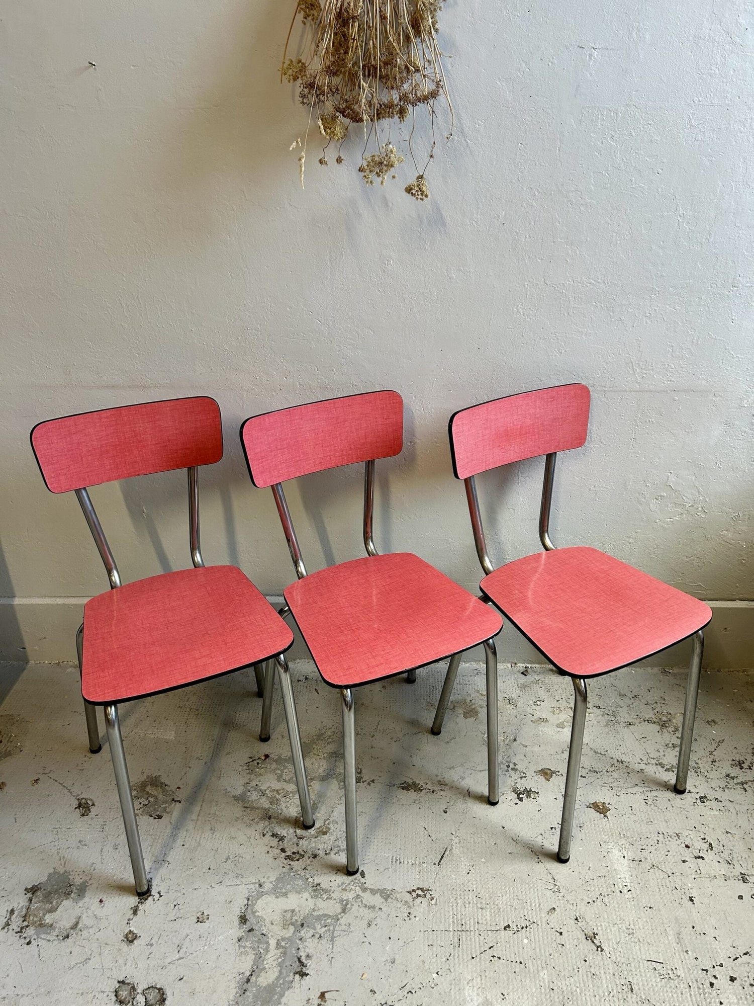 Trois chaises vintage en formica rose corail et piétement en métal chromé tubulaire, alignées contre un mur gris clair. Au-dessus d'elles, un bouquet suspendu de fleurs séchées apporte une touche poétique à l'ensemble.