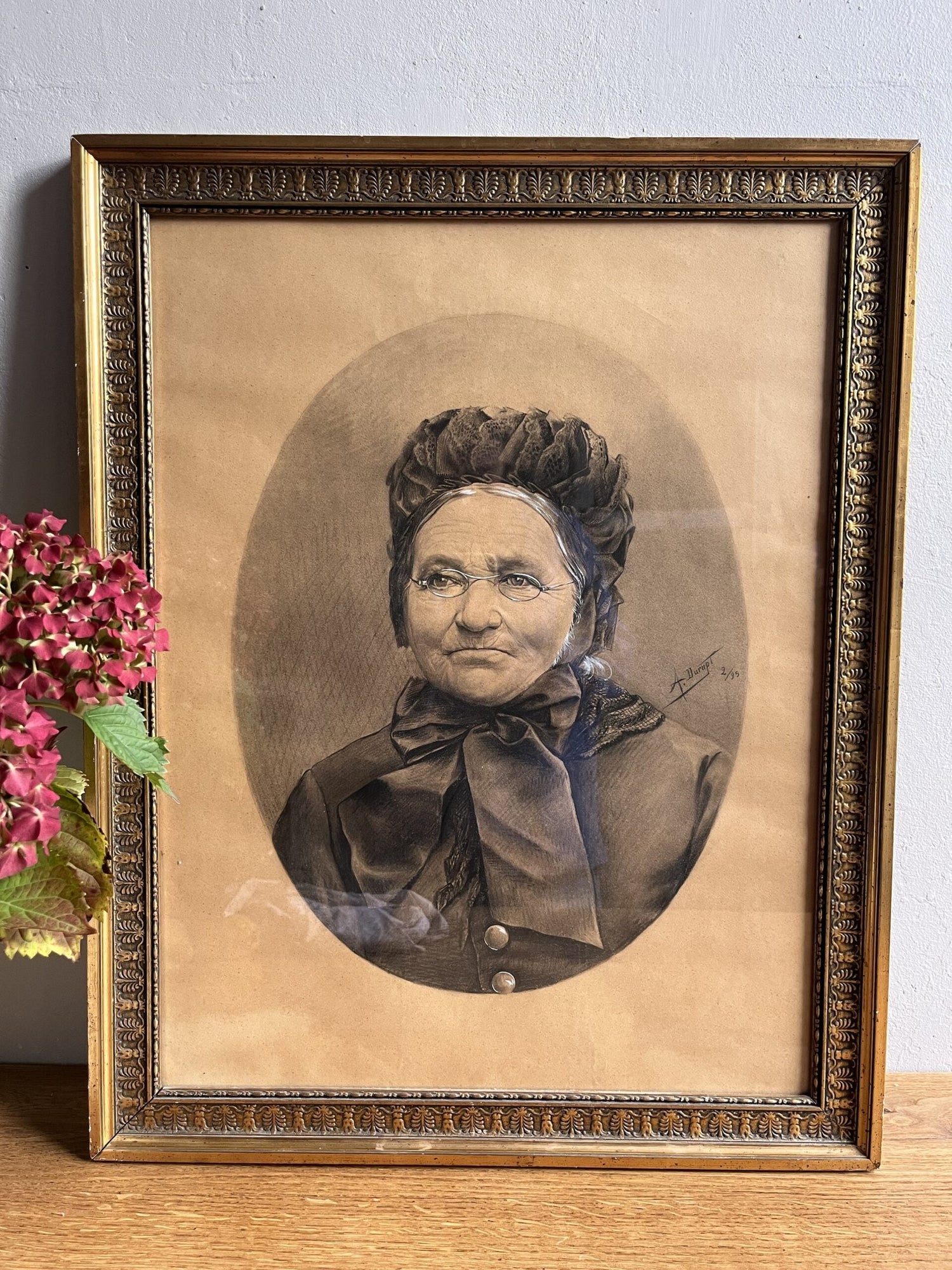 Portrait ancien au fusain ou dessin d'une femme âgée à lunettes et coiffe, de forme ovale sur fond beige. Présenté sous verre dans un grand cadre en bois doré mouluré et sculpté, posé sur un meuble en bois à côté d'hortensias roses.