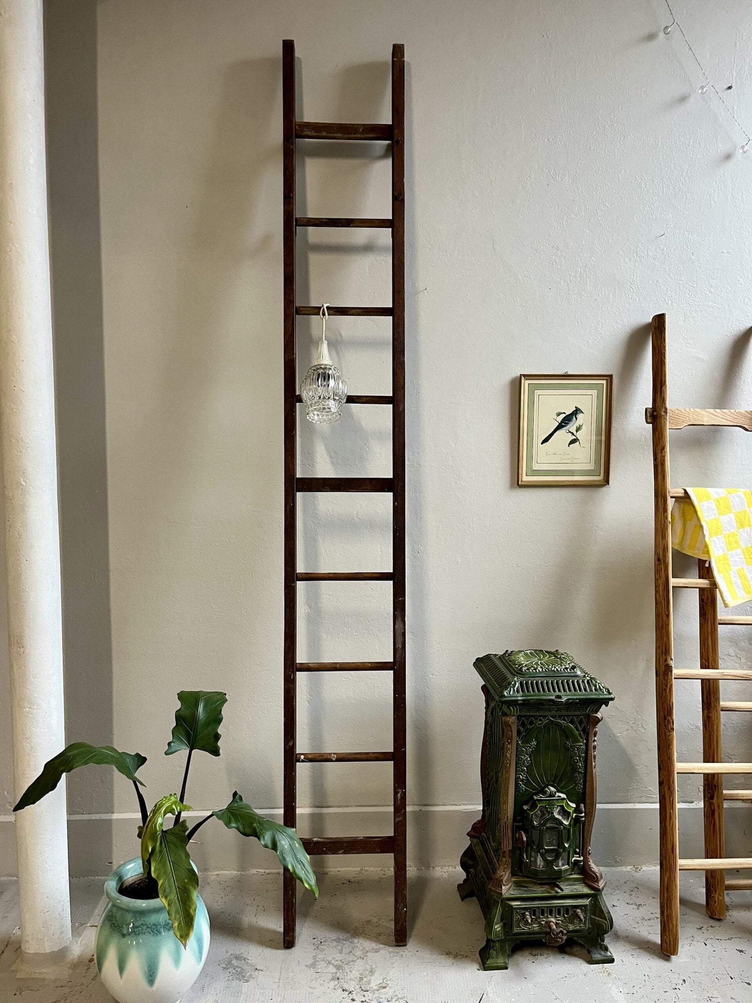 Grande échelle de meunier ancienne en bois foncé posée contre un mur blanc, décorée d'une suspension en verre ciselé. Au sol, une plante verte dans un pot en céramique turquoise et un poêle en fonte émaillée verte, à côté d'un cadre oiseau vintage.