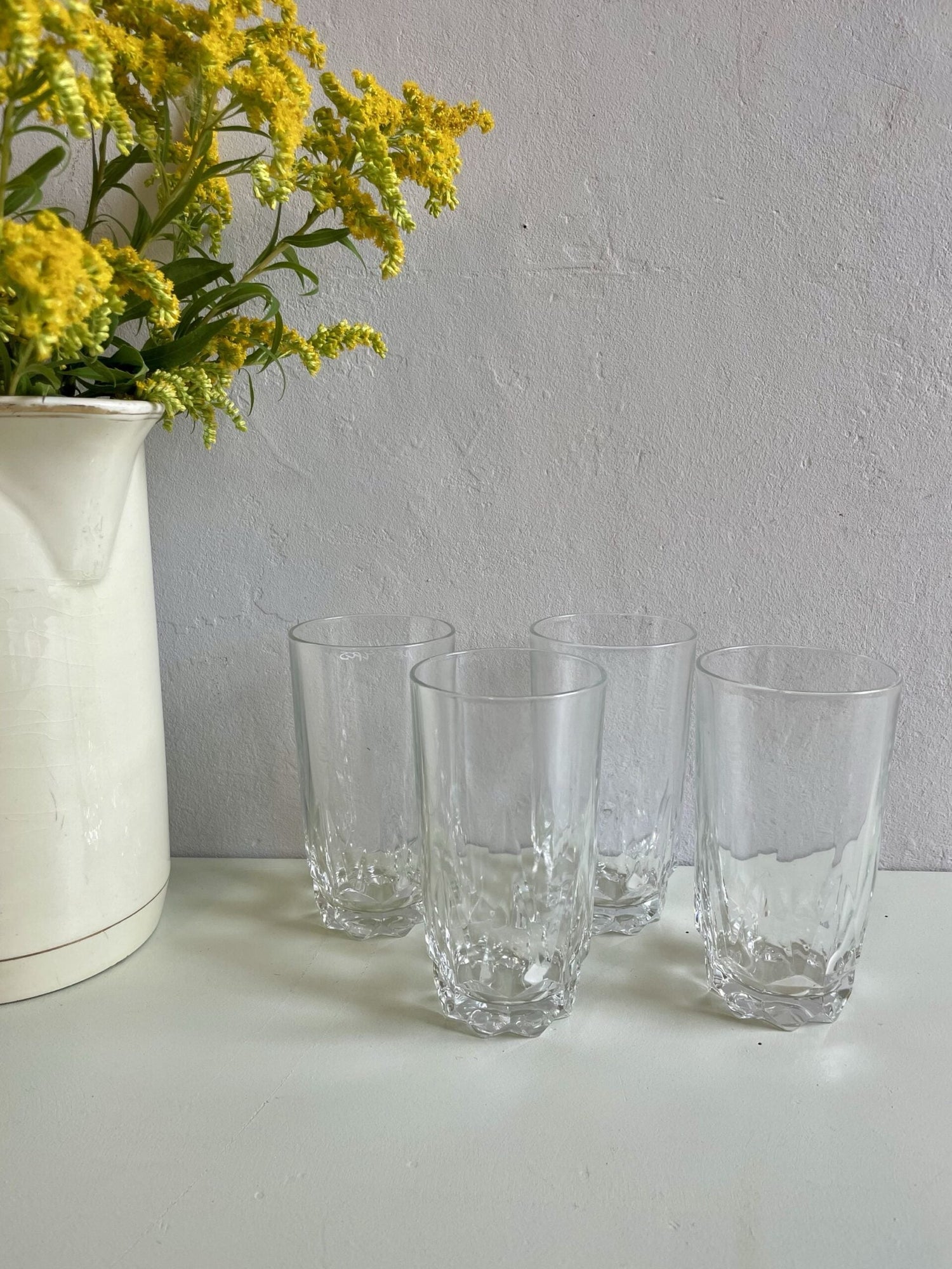 4 verres à eau en verre transparent sur un plateau vert pâle et un fond gris. A gauche, un pichet blanc avec du mimosa