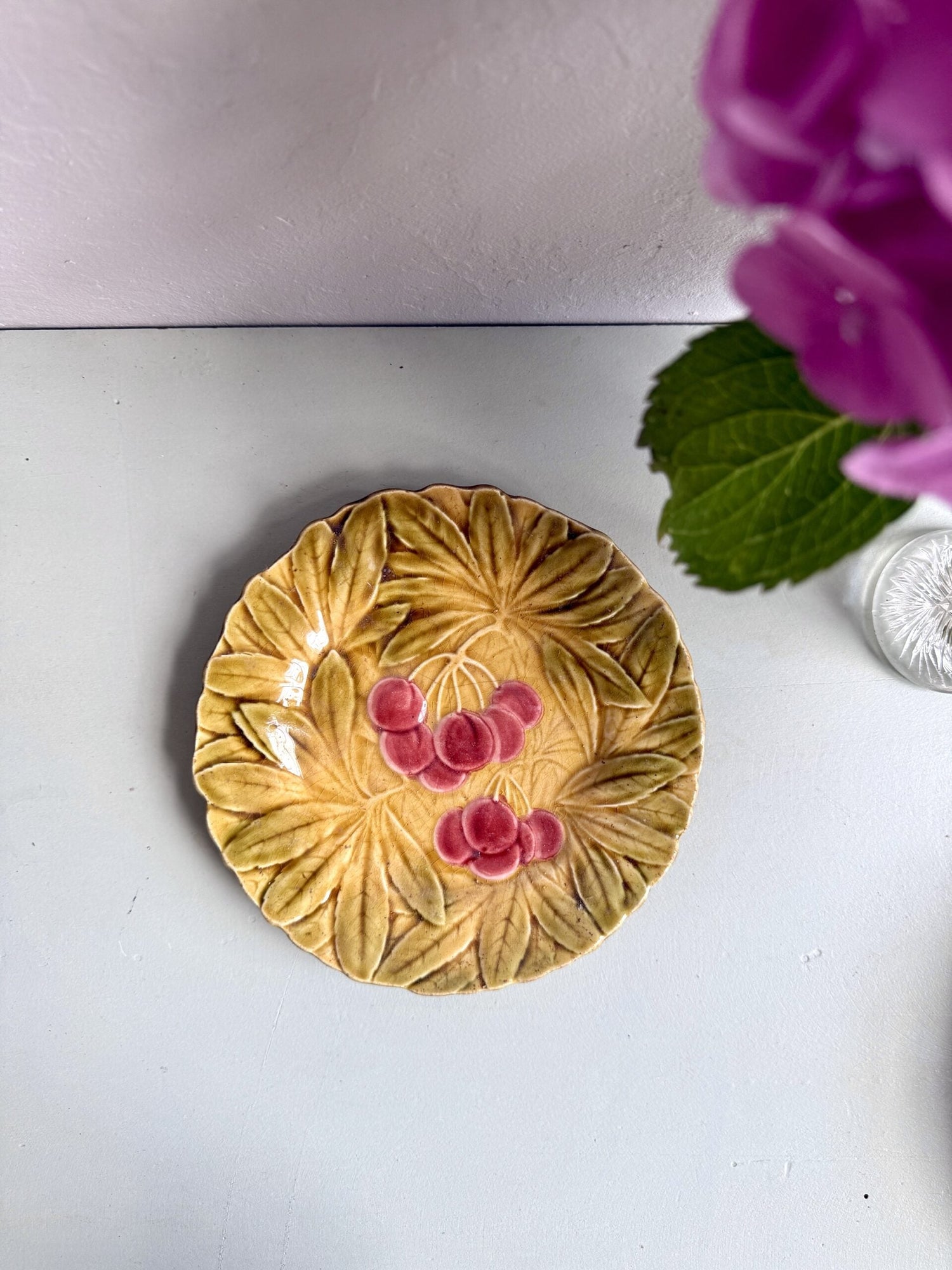 Assiette à dessert ancienne en barbotine à décor en relief de cerises roses et de grandes feuilles dans les tons ocre et vert. Vue du dessus, posée sur un fond blanc uni avec une fleur d'hortensia rose vif visible au premier plan à droite.