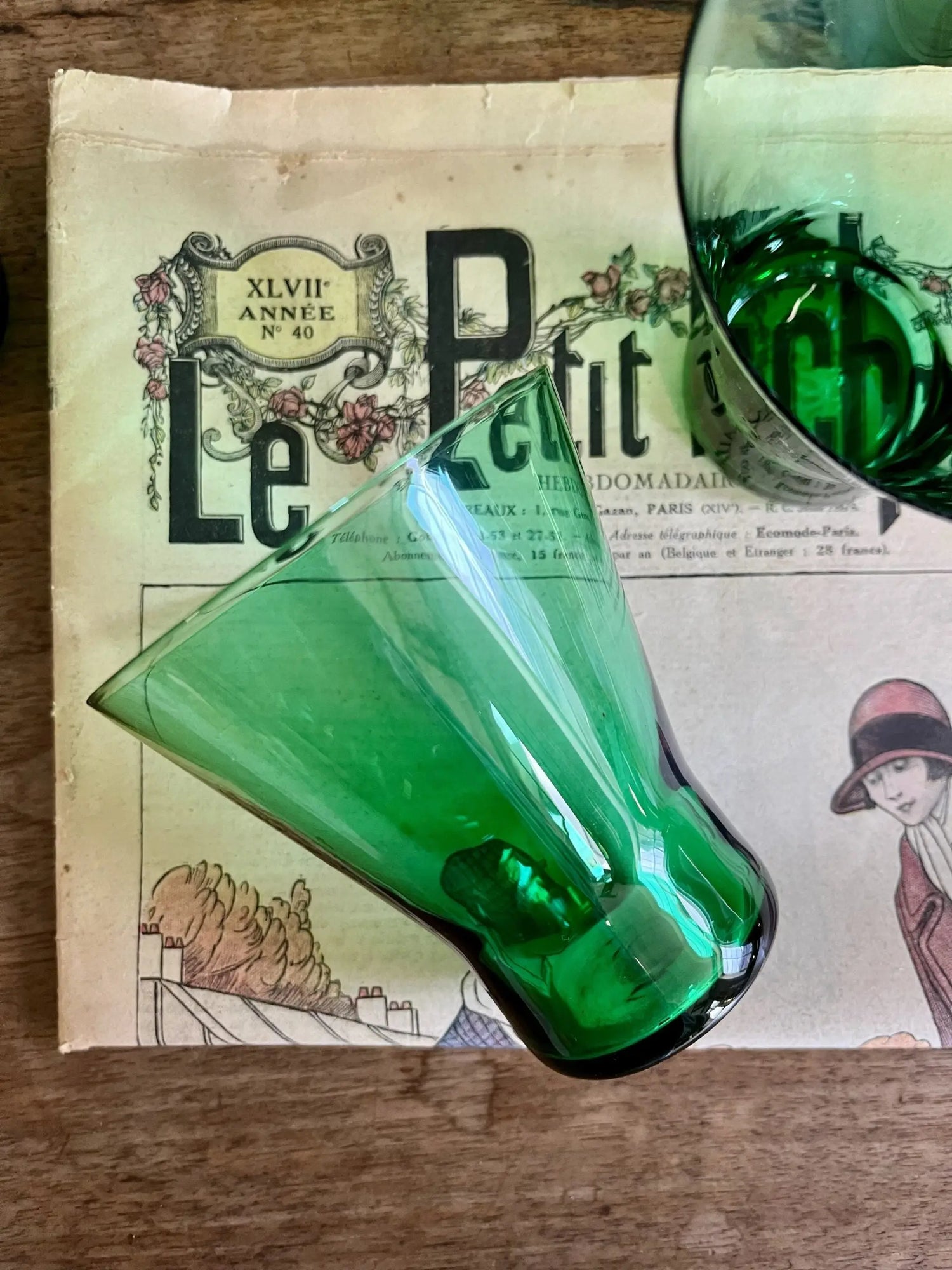 Deux verres anciens en verre soufflé de couleur vert émeraude, dont un couché au premier plan, posés sur un ancien journal illustré rétro intitulé "Le Petit Écho de la Mode", le tout disposé sur une table en bois brut.