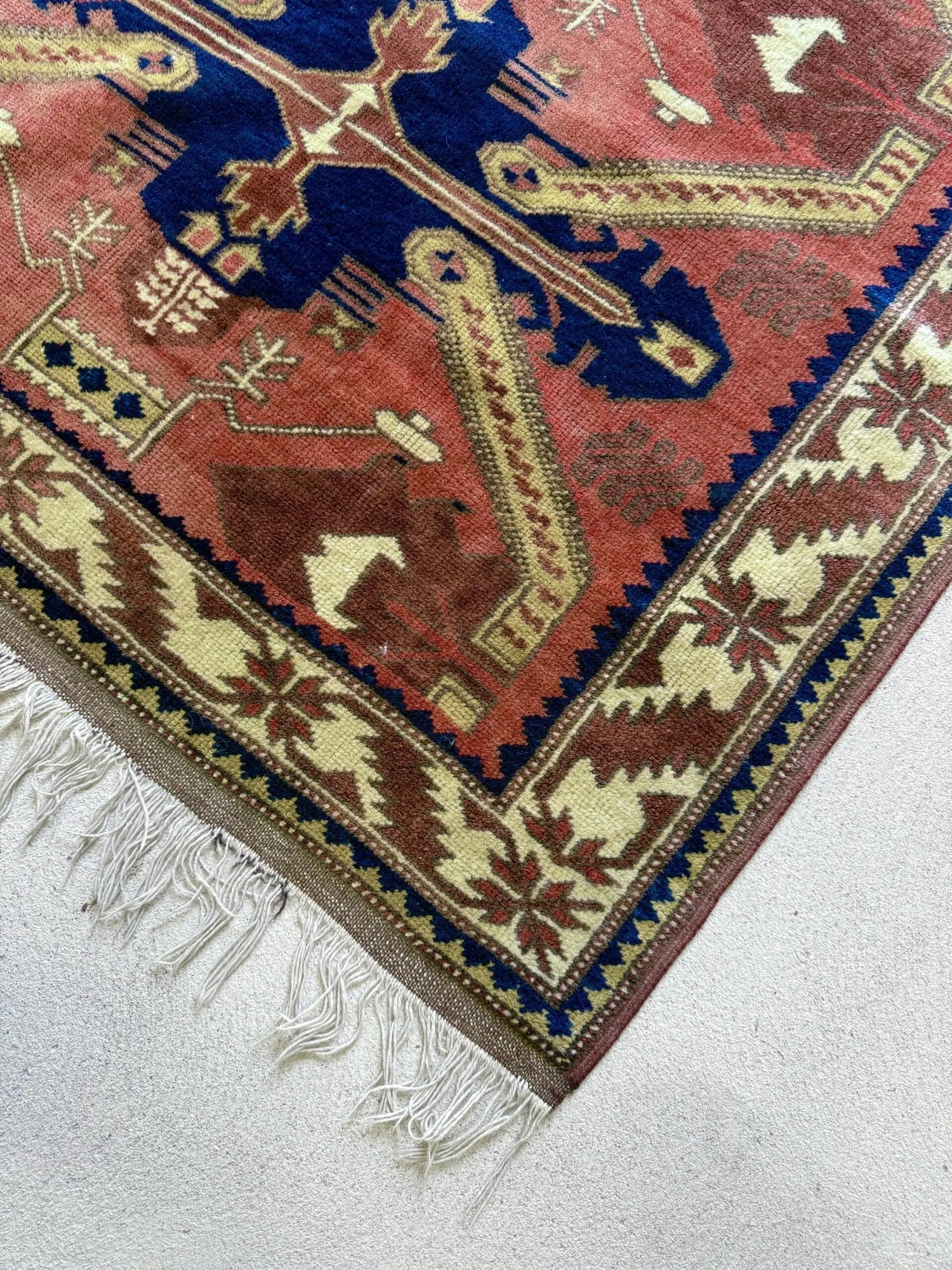 Tapis ancien en laine de style persan ou kilim à motifs géométriques, combinant des teintes rouge brique, bleu cobalt, beige et marron. Détail du coin avec franges blanches en coton, présenté à plat sur un sol clair.