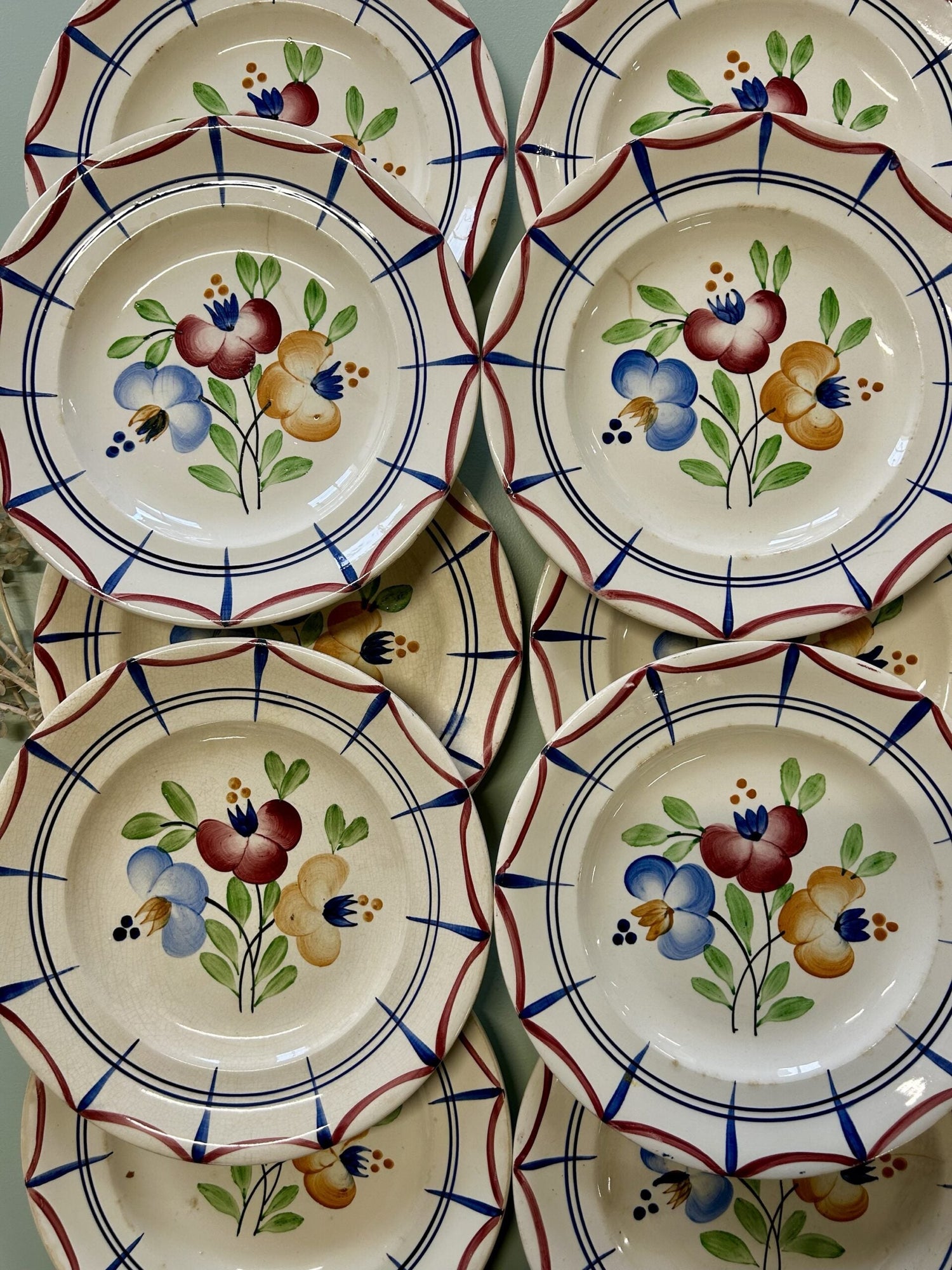Vue du dessus sur des assiettes empilées. Au centre de l'assiette un bouquet floral bleu rouge jaune. Sur le pourtour, des motifs géométrique dans les même teintes que le bouquet: bleu et bordeaux