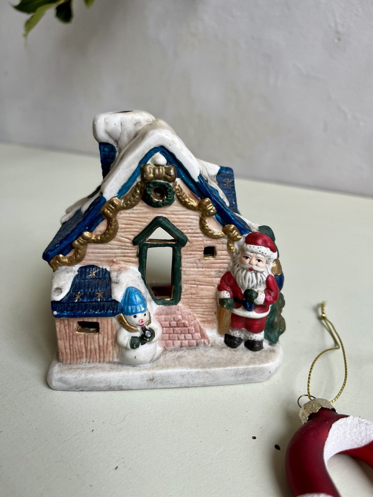 Petite maison de Noël vintage en céramique ou photophore, décorée d'une figurine de Père Noël et d'un bonhomme de neige sous un toit enneigé bleu et blanc. Posée sur une surface claire à côté d'une suspension de Noël rouge avec cordon doré.