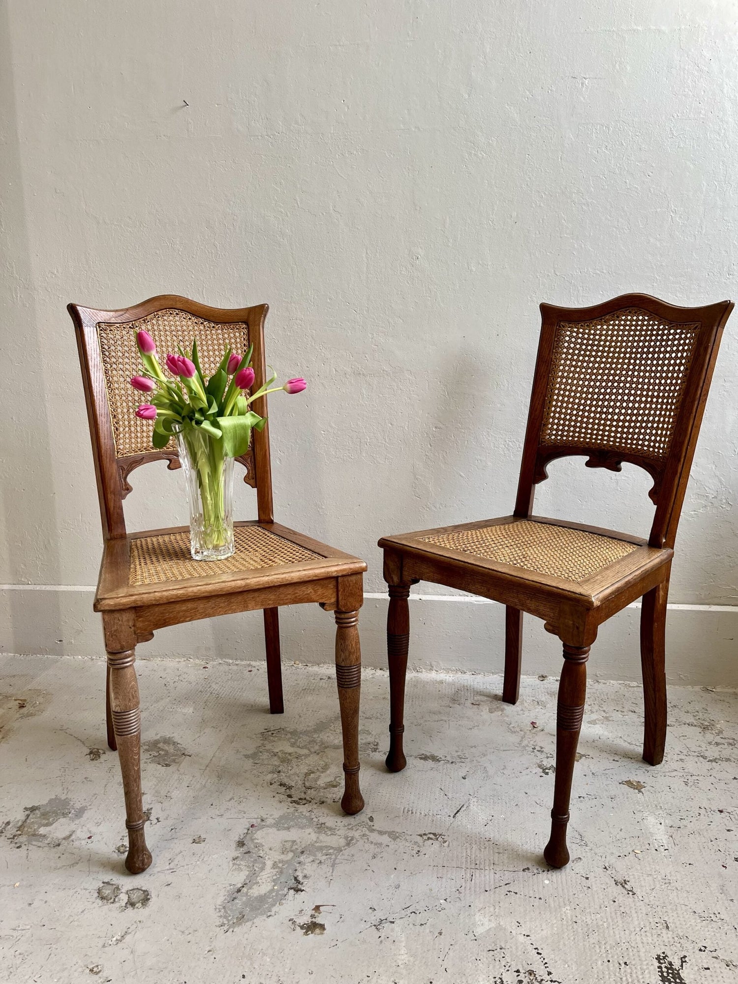 Paire de chaises anciennes en bois et cannage (dossier et assise), de style Louis XVI ou Directoire à pieds tournés, posées sur un sol brut contre un mur clair. La chaise de gauche porte un vase en verre transparent garni de tulipes roses.