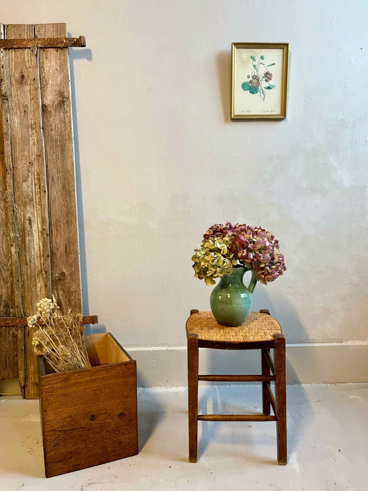 Tabouret rustique en bois à assise paillée, surmonté d'un pichet vert en céramique garni d'hortensias séchés. À gauche, un bac en bois remplit de fleurs séchées repose devant un panneau ancien. Un petit cadre de botanique décore le mur blanc.