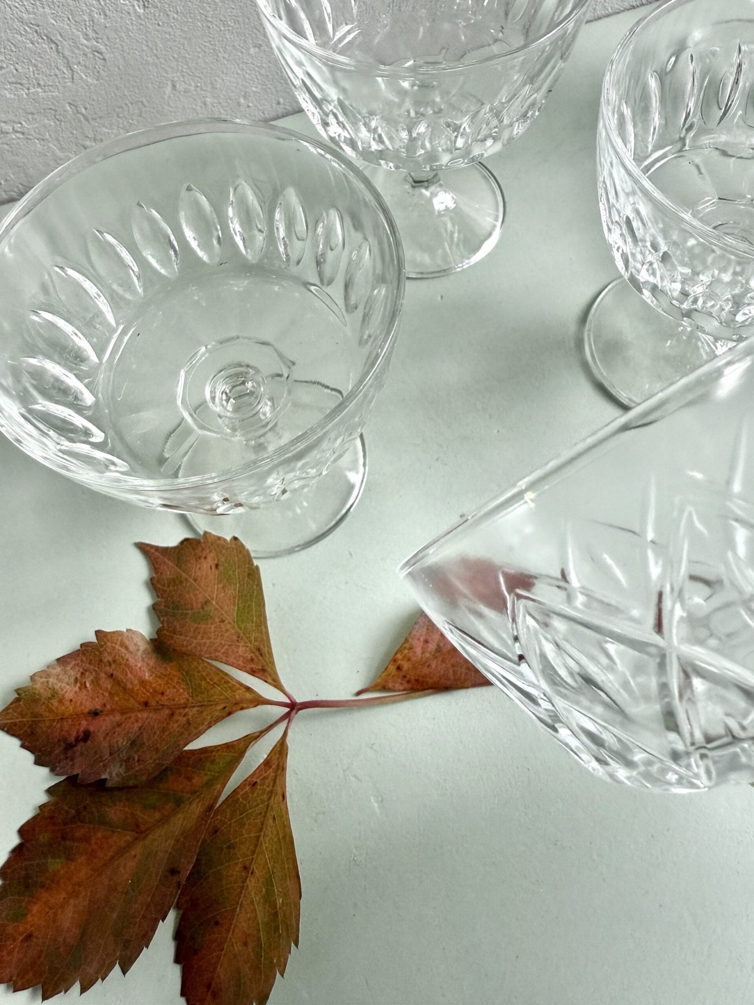 Coupes à champagne anciennes en verre moulé transparent à reliefs géométriques, sur fond clair. Une feuille de vigne vierge aux tons automnaux rousses est posée au premier plan, ajoutant une touche de saison.