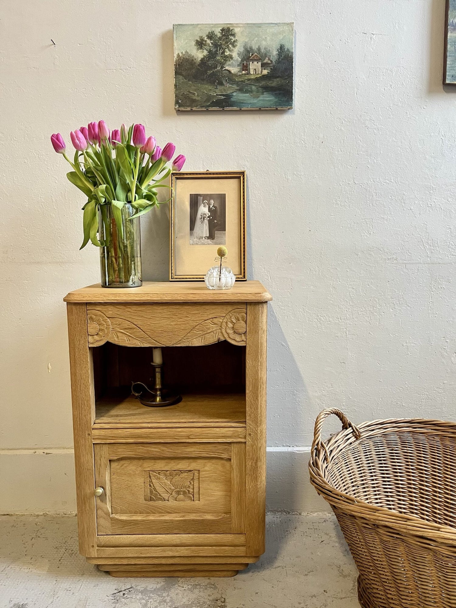 Chevet art deco avec tiroir grave niche contenant un chandelier. Ambiamce déco avec un bouquet de tulipe rose et 2 cadres. une panière en rotin sur la droite 