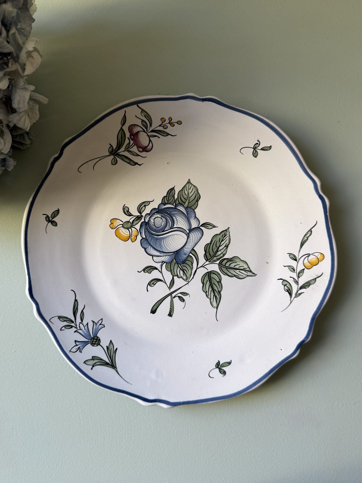 Assiette ancienne en faïence à bord festonné souligné de bleu, décorée d'une grande rose bleue centrale peinte à la main et de motifs floraux. Posée sur un fond vert clair, elle est accompagnée en haut à gauche de fleurs d'hortensia séchées.