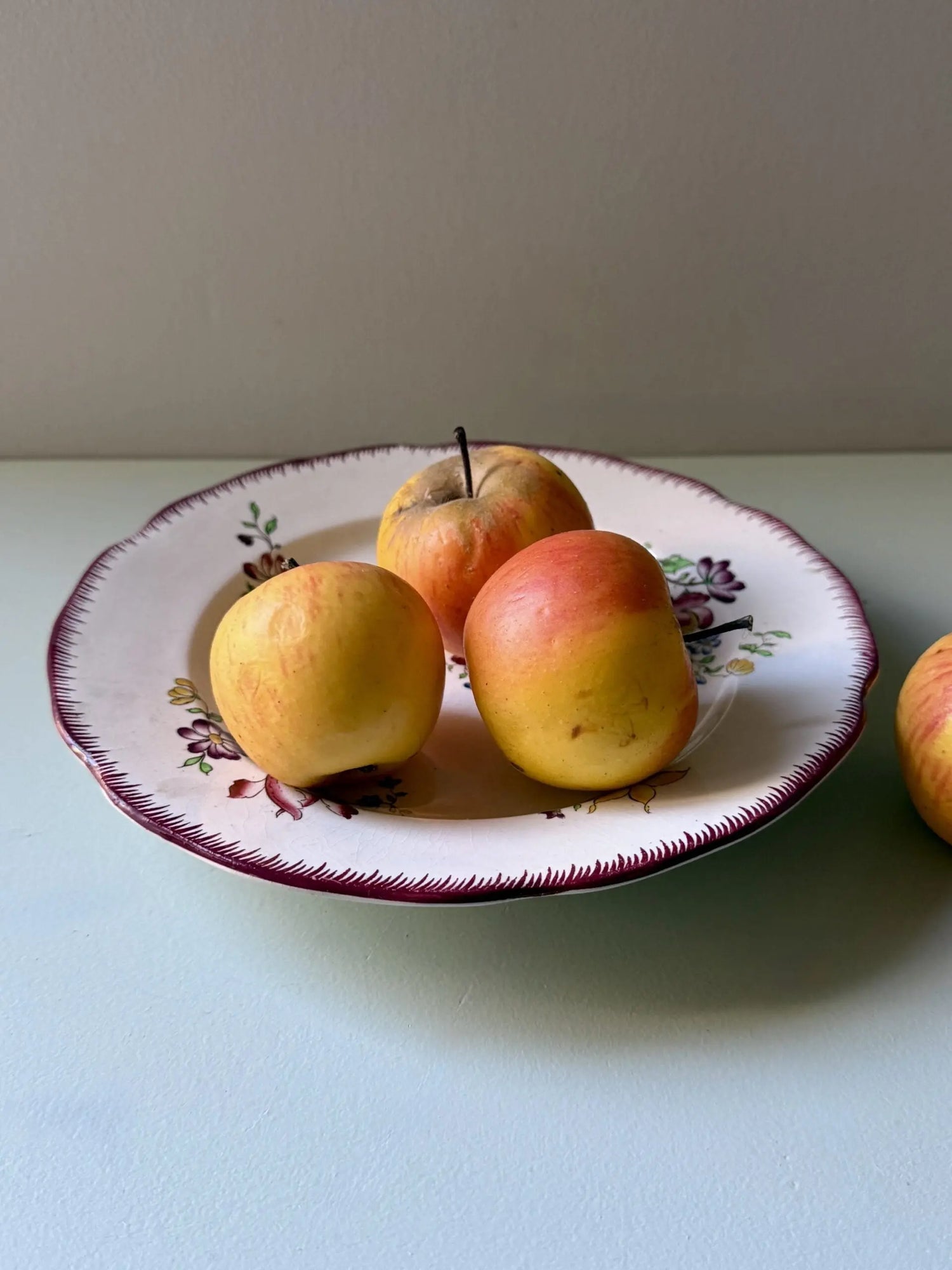 Assiette creuse ou jatte ancienne en faïence blanche à bord peigné bordeaux, décorée de motifs floraux discrets. Elle contient trois pommes anciennes jaunes et rouges, posée sur un fond uni vert amande à côté d'une quatrième pomme.