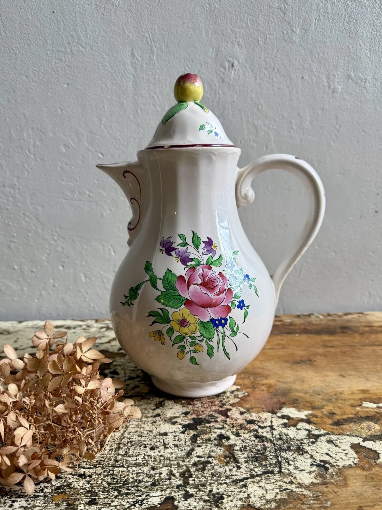 Cafetière ou verseuse ancienne en faïence blanche à côtes, décorée d'un bouquet de roses et fleurs peintes. Couvercle surmonté d'un bouton en forme de fruit. Posée sur un meuble en bois patiné à côté d'hortensias séchés.