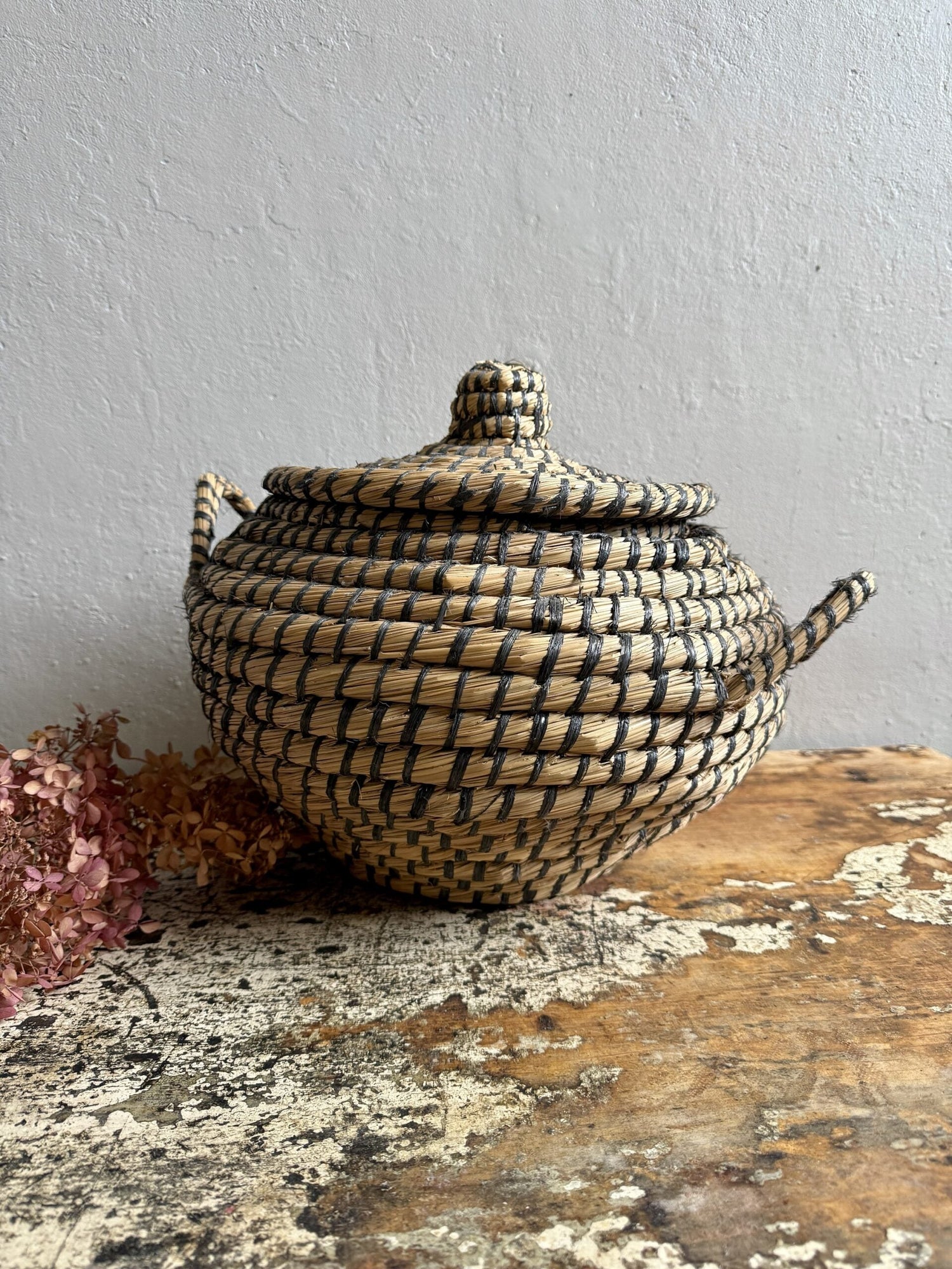 Pot ou panier en paille tressée naturelle de style berbère ou bohème, avec un motif quadrillé noir, un couvercle avec poignée et une anse latérale, posé sur une surface en bois usé avec des fleurs séchées.