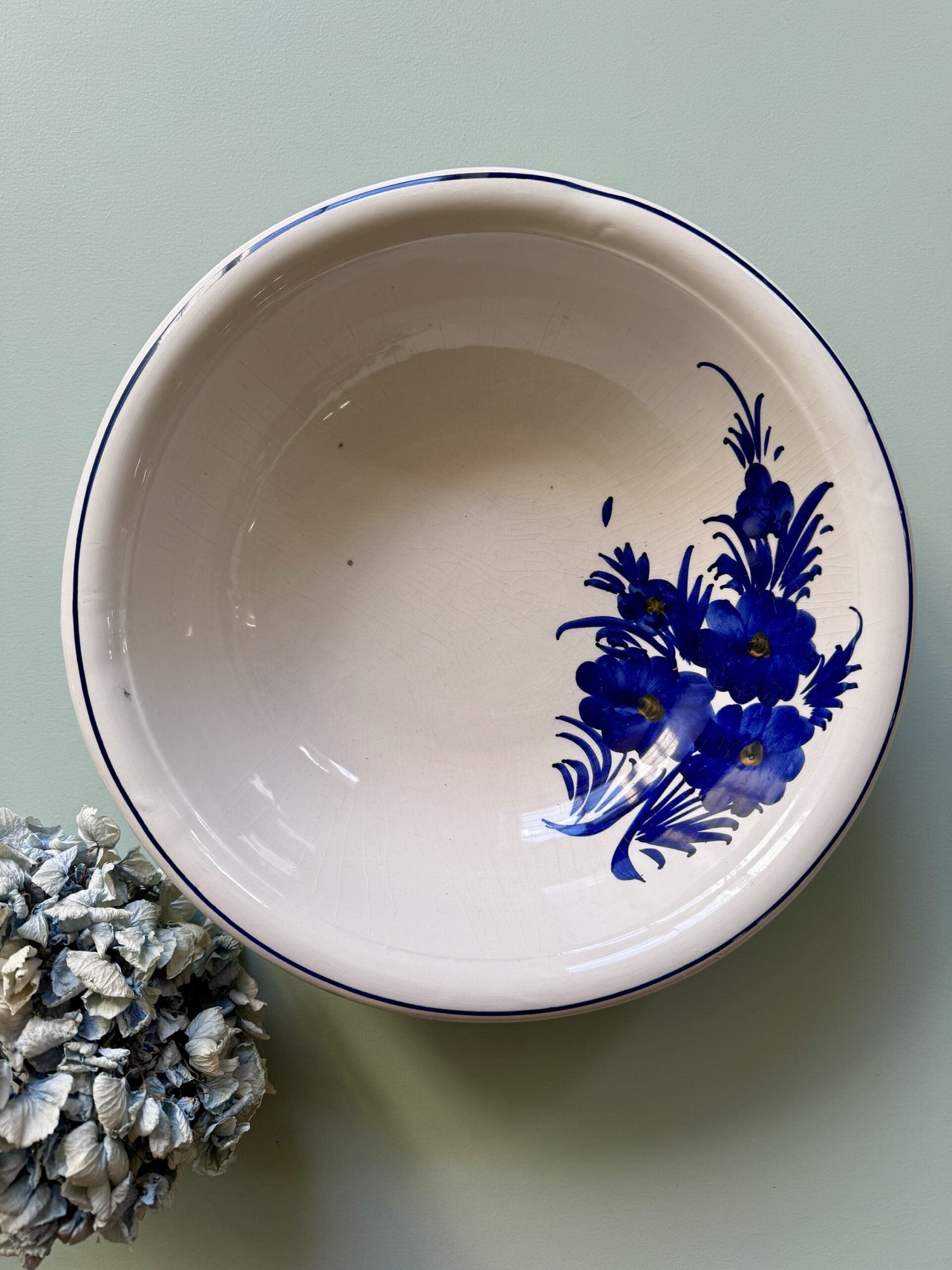 Saladier terre de fer blanc avec liseré bleu et décor floral bleu foncé sur le côté. Posé sur un fond vert clair, avec une hortensia séché bleu sur le côté gauche