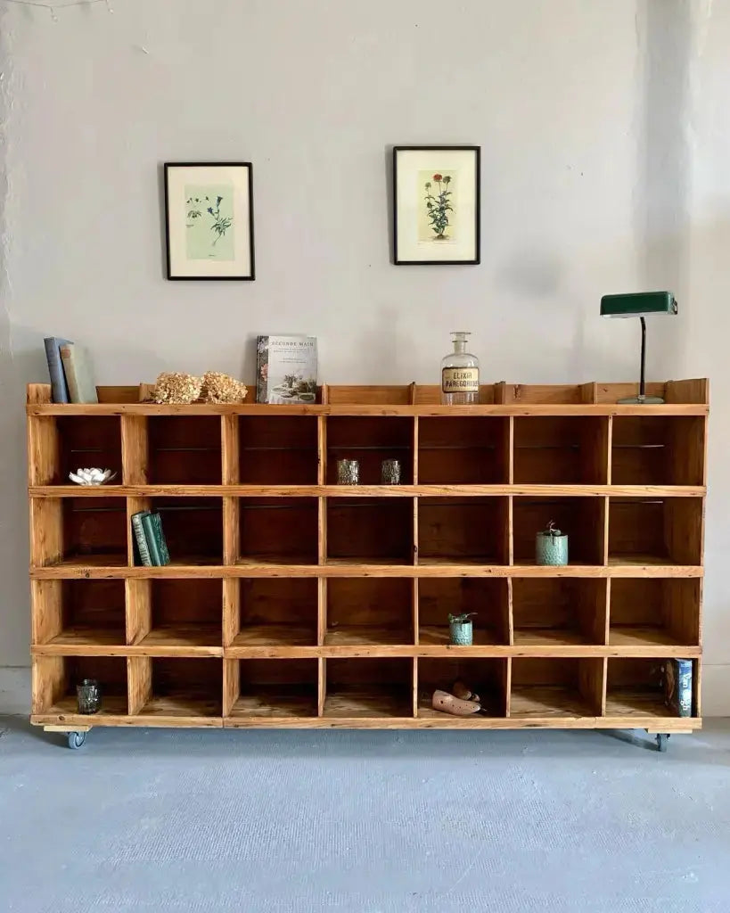 Grand meuble de métier ou casier de tri ancien en bois brut monté sur roulettes. Ses compartiments accueillent des livres, de la verrerie et de petits pots turquoises. Dessus : flacon d'apothicaire, lampe de notaire verte et cadres botaniques au mur.