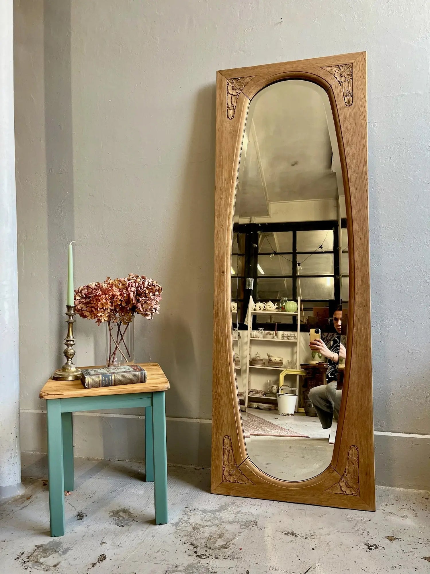 Grand miroir psyché ancien en bois clair sculpté de fleurs de style Art Déco ou Art Nouveau, posé au sol contre un mur gris à côté d'un tabouret turquoise portant un vase d'hortensias séchés, un livre et un bougeoir.