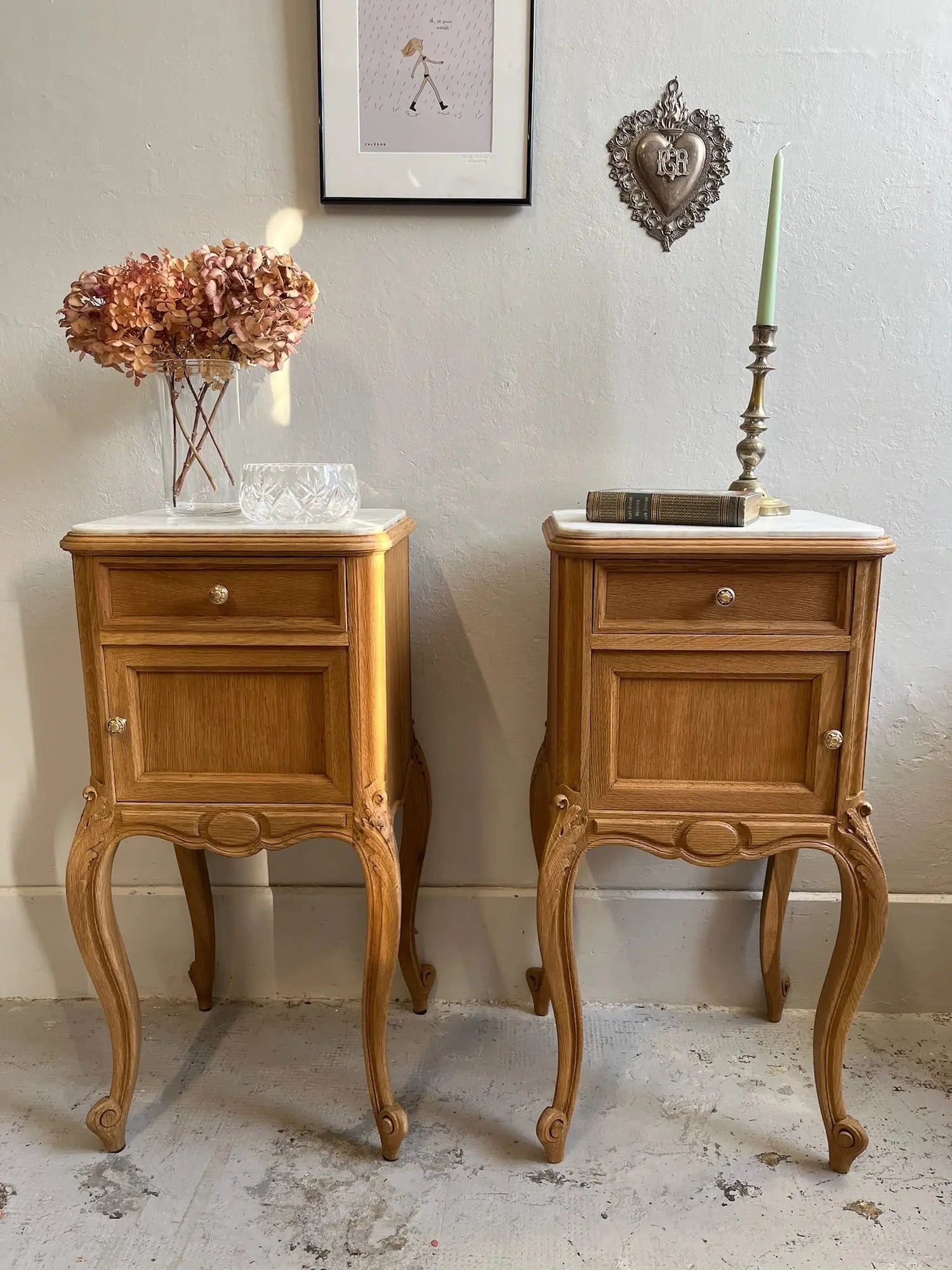 Paire de tables de chevet anciennes en bois clair de style Louis XV, avec pieds galbés et plateau en marbre blanc. Dessus posés : vase d'hortensias séchés, coupelle en verre, livre ancien et bougeoir. Au mur : un ex-voto cœur et un cadre.