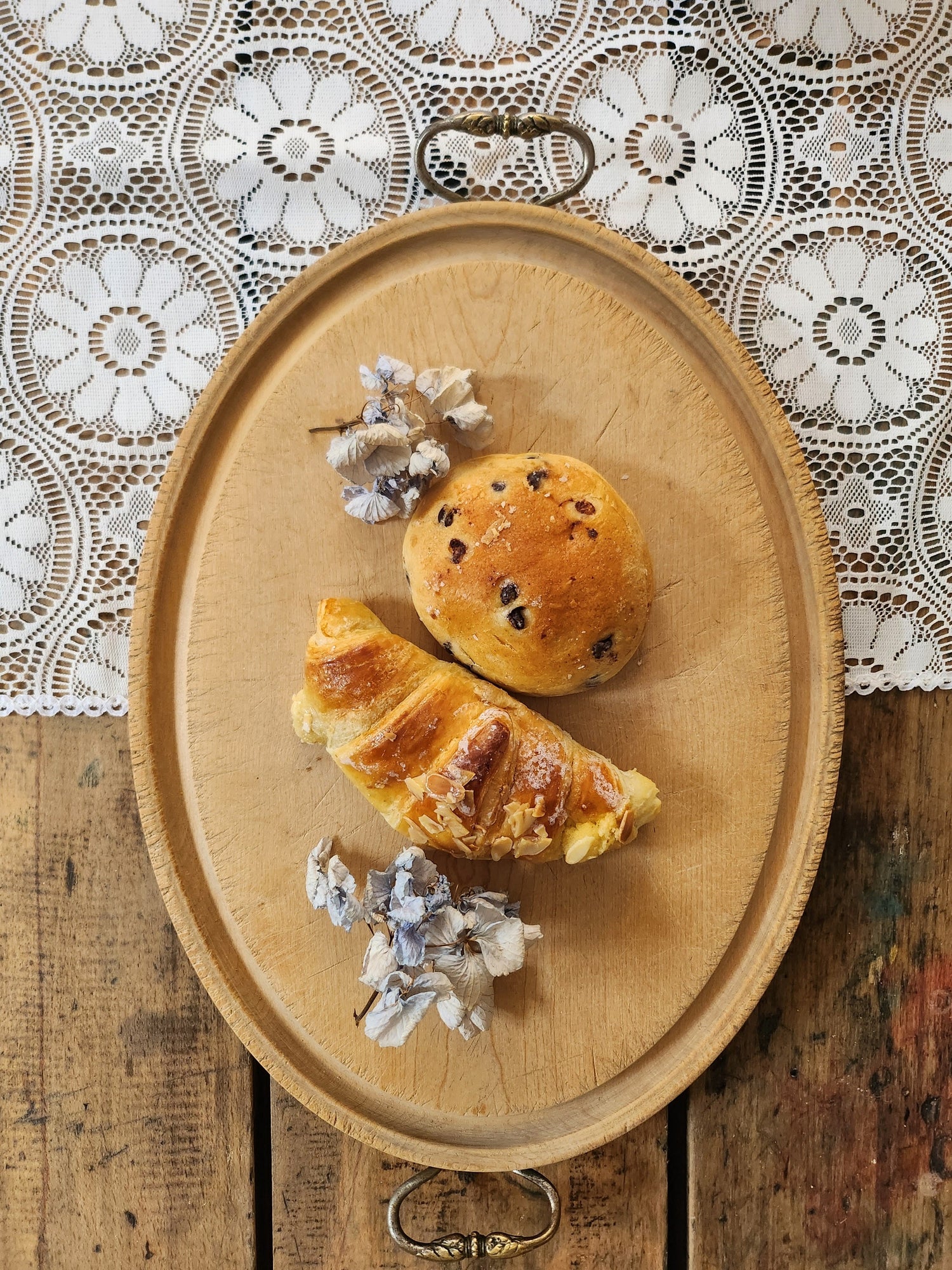 Plateau ovale ancien en bois clair avec deux poignées en métal ciselé, utilisé comme plateau de présentation. Dessus posés : un croissant aux amandes, un petit pain aux pépites de chocolat et des fleurs séchées, sur fond de nappe en dentelle blanche.