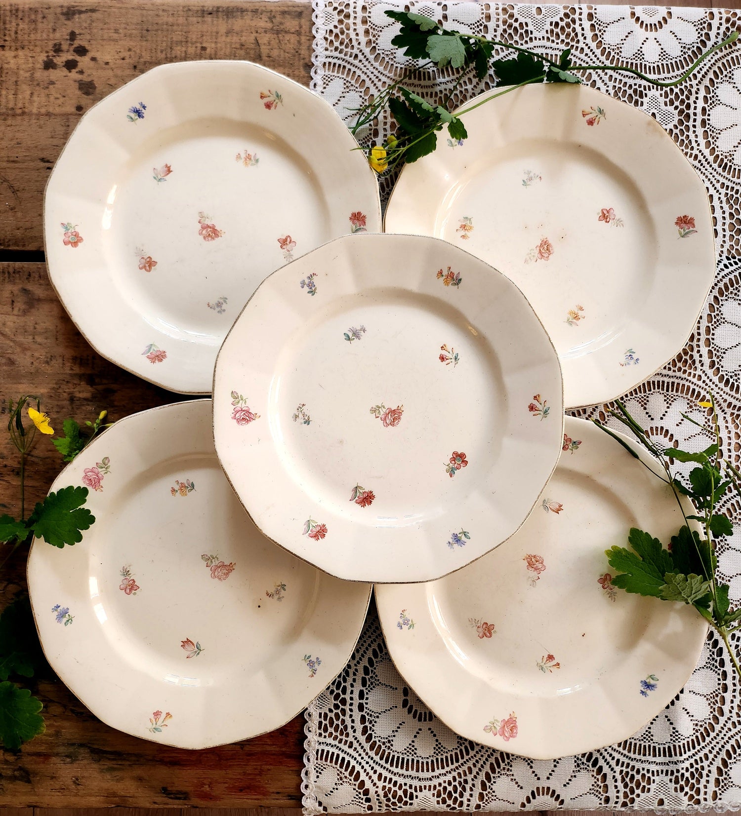 5 assiettes en céramique blanche avec décor de fleurs roses et bleues sur fond bois et dentelle. Des fleurs jaunes viennent décorer.