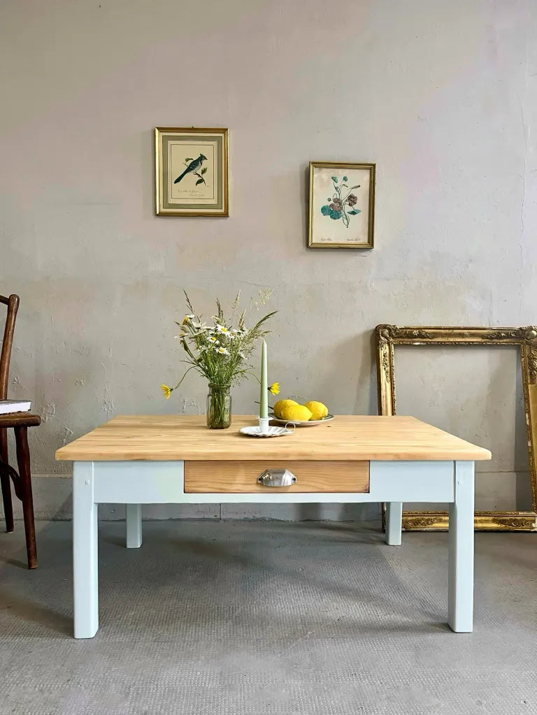 Table basse en bois au piètement peint en bleu clair, dotée d'un tiroir central. Sur le plateau : bouquet de marguerites, bougie et citrons. À l'arrière, deux petits cadres botaniques et un grand cadre doré au sol décorent le mur gris.