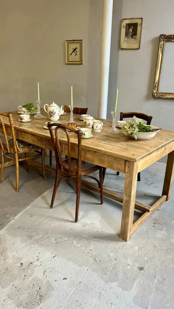 Grande table de ferme ancienne en bois brut entourée de chaises dépareillées (style Baumann ou Thonet). Dessus dressé : service à thé ou café en faïence fleurie, bougeoirs avec bougies vertes et coupes de fleurs. Au mur gris, des cadres vintage.