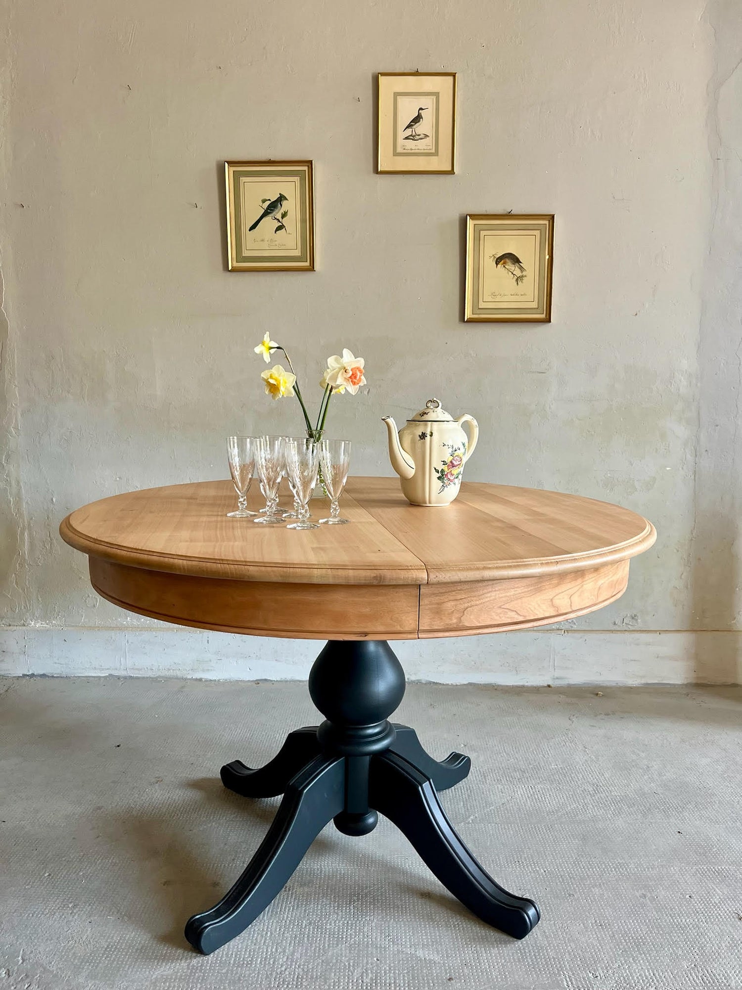 Table ronde en bois au piètement central noir, surmontée de flûtes en cristal, d'une cafetière fleurie et de jonquilles. Au mur gris, trois petits cadres anciens illustrant des oiseaux complètent cette ambiance brocante raffinée.
