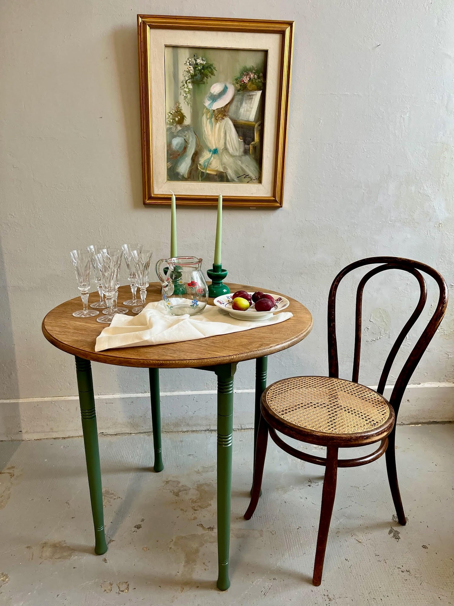 Table ronde en bois aux pieds verts, dressée avec flûtes en cristal, pichet fleuri, bougies et prunes. À droite, une chaise bistrot en cannage. Un tableau classique d'une jeune fille au piano orne le mur gris en arrière-plan.
